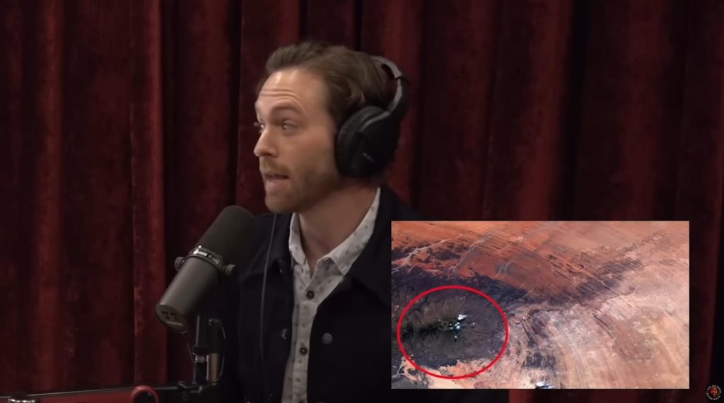screen capture of a conspiracy theorist in a dorky button up shirt and jacket wearing headphones with mouth slight agape speaking nonsense into a large microphone owned by joe rogan next to a screen shot of the Richat Structure